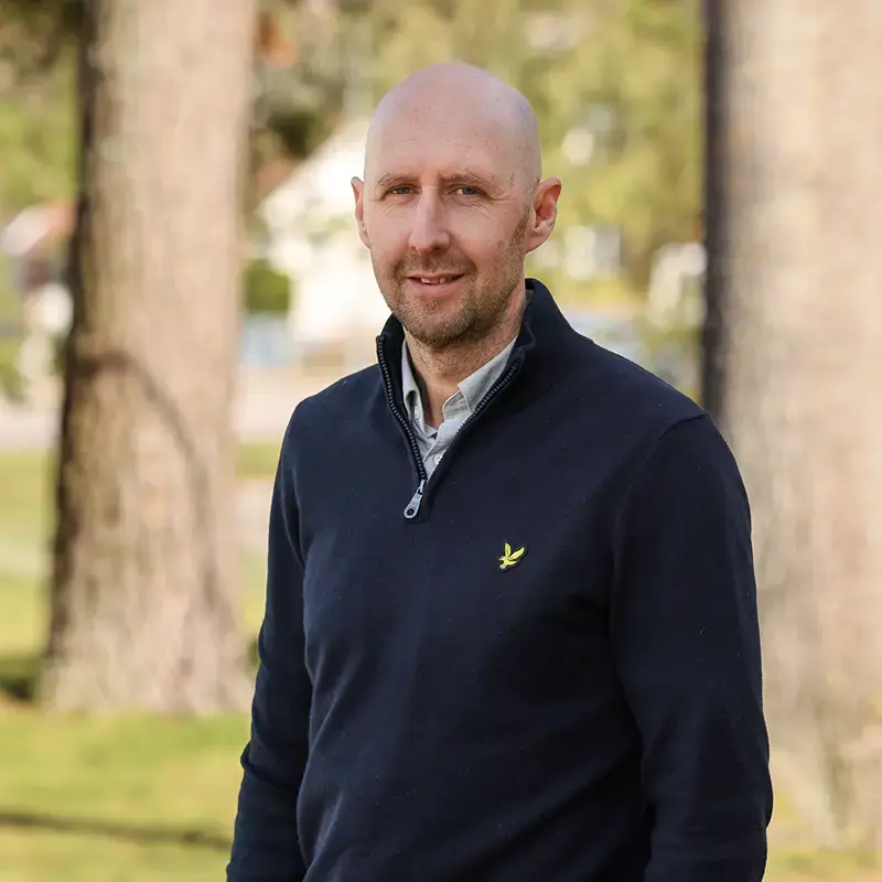 Man tittar in i kameran. I bakgrunden natur med träd och grön gräsmatta.